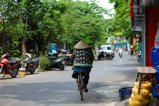 A person wearing a traditional hat rides a bicycle through a bustling city street lined with motorcycles.