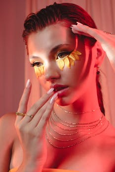 Stylish portrait of a woman with artistic golden eye makeup and elegant jewelry in a warm, indoor setting.