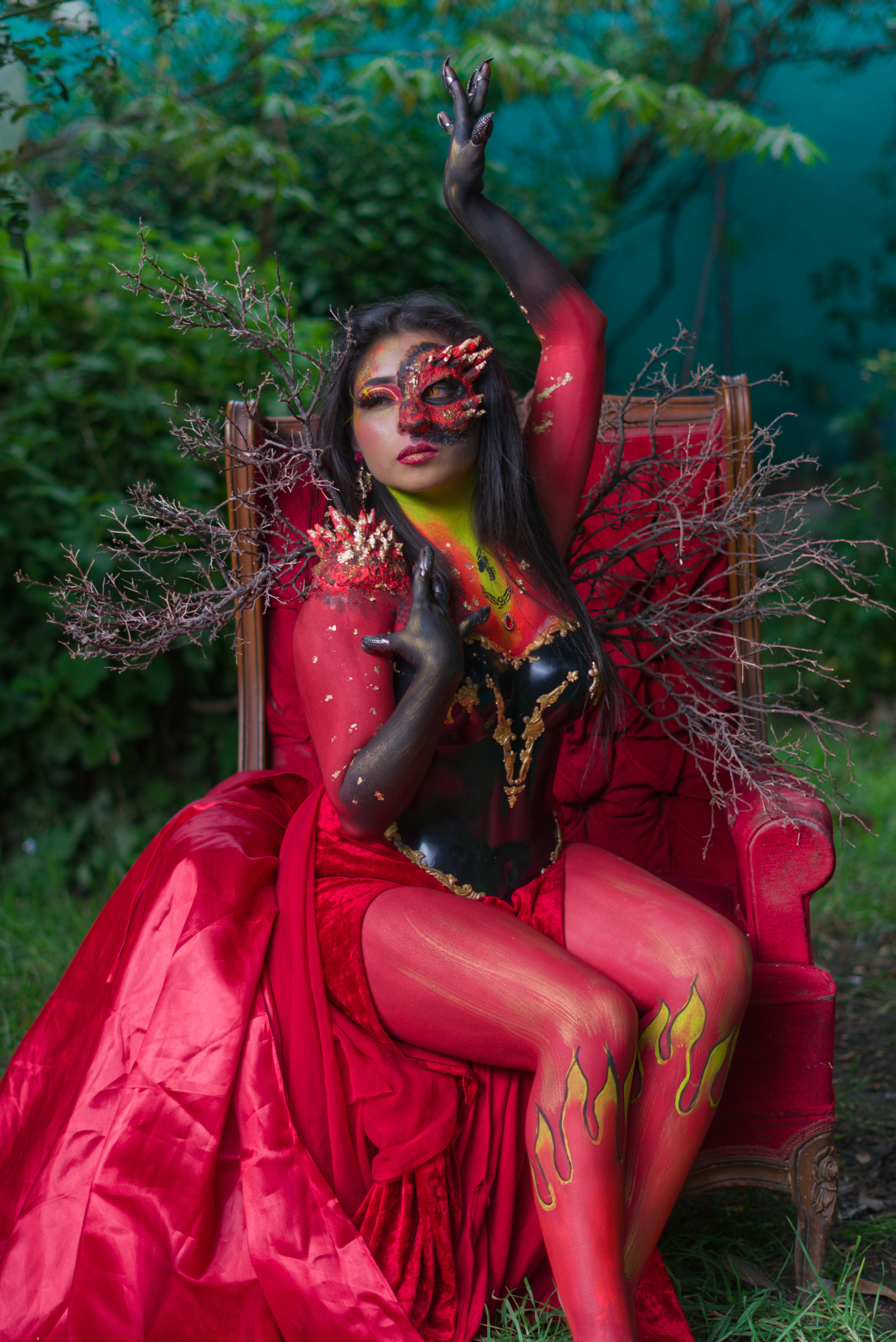 Free A striking woman in vivid costume and body paint poses on a red chair with dramatic makeup and natural elements. Stock Photo
