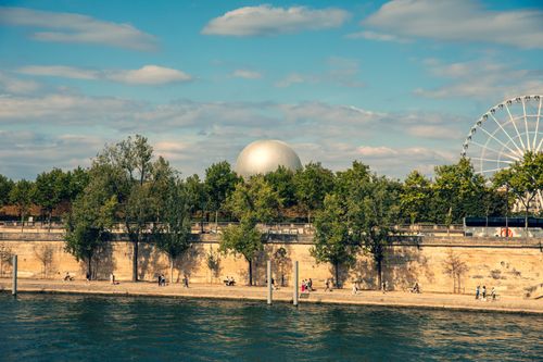 Free Picturesque view of a sunny Paris riverbank with trees, a Ferris wheel, and dome. Stock Photo