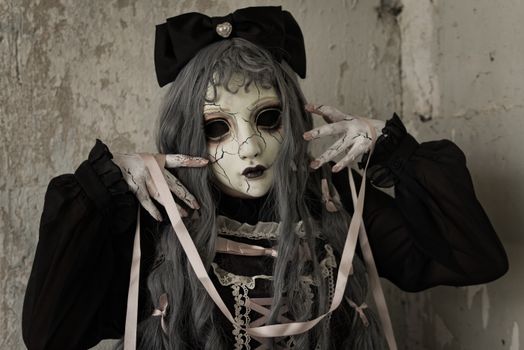 A person in eerie doll-like cosplay with a cracked mask, ribbons, and dark clothing, indoors with dark mood lighting.