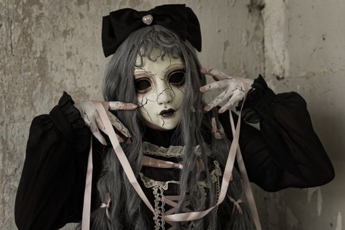 Free A person in eerie doll-like cosplay with a cracked mask, ribbons, and dark clothing, indoors with dark mood lighting. Stock Photo