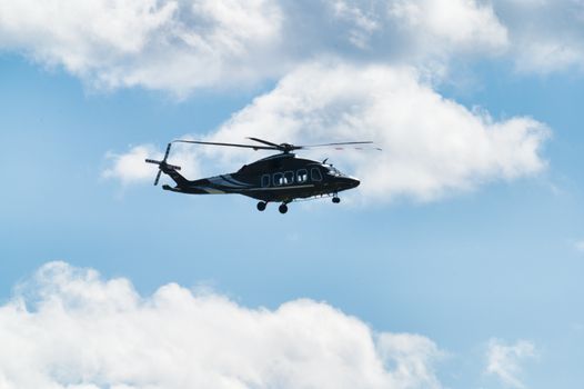 A sleek helicopter flying high against a vibrant blue sky with fluffy clouds, captured above Poprad, Slovakia.