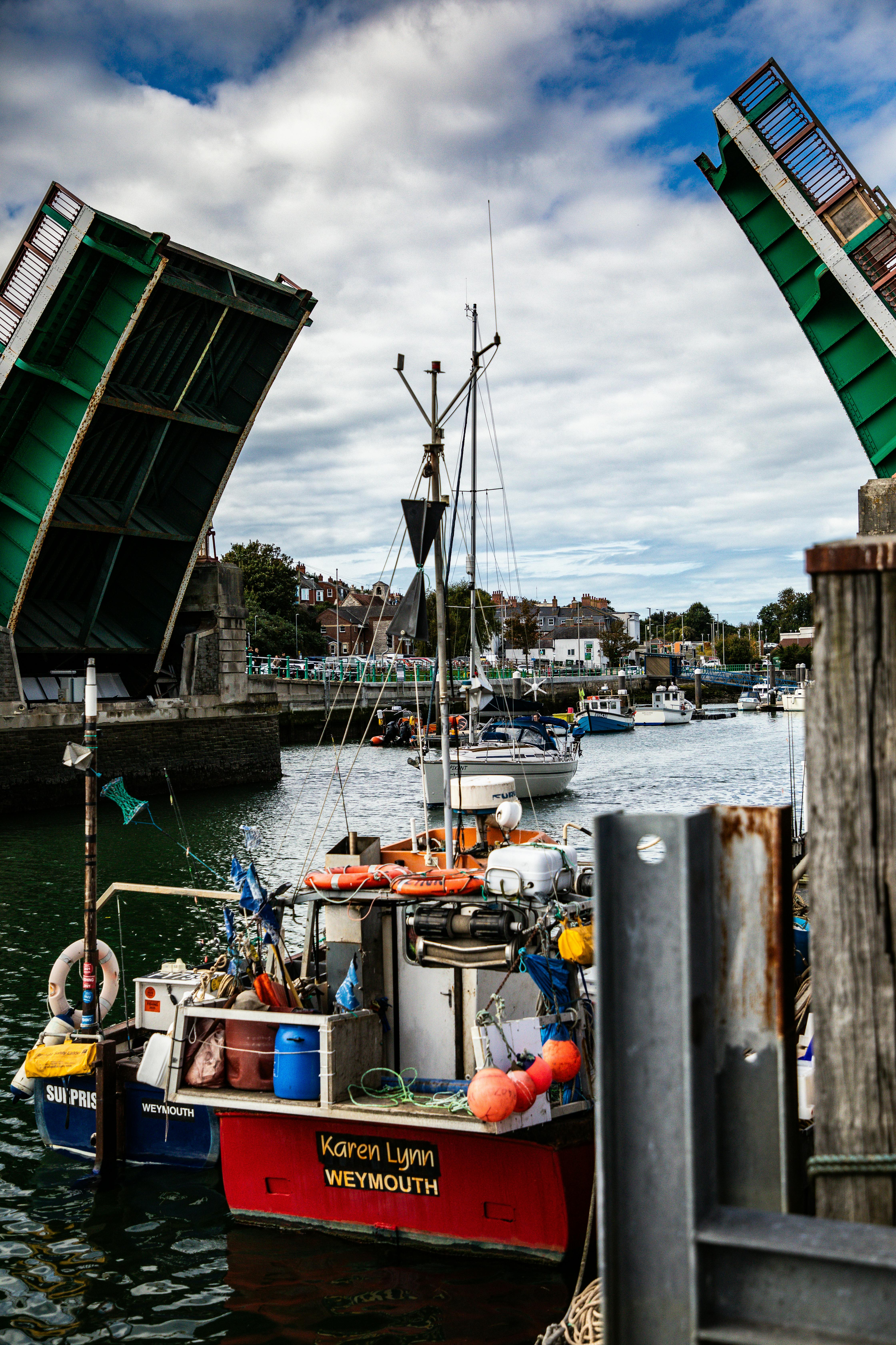 Fishing boats passing under Weymouth Town Bridge with vibrant colors and a bustling harbor scene.