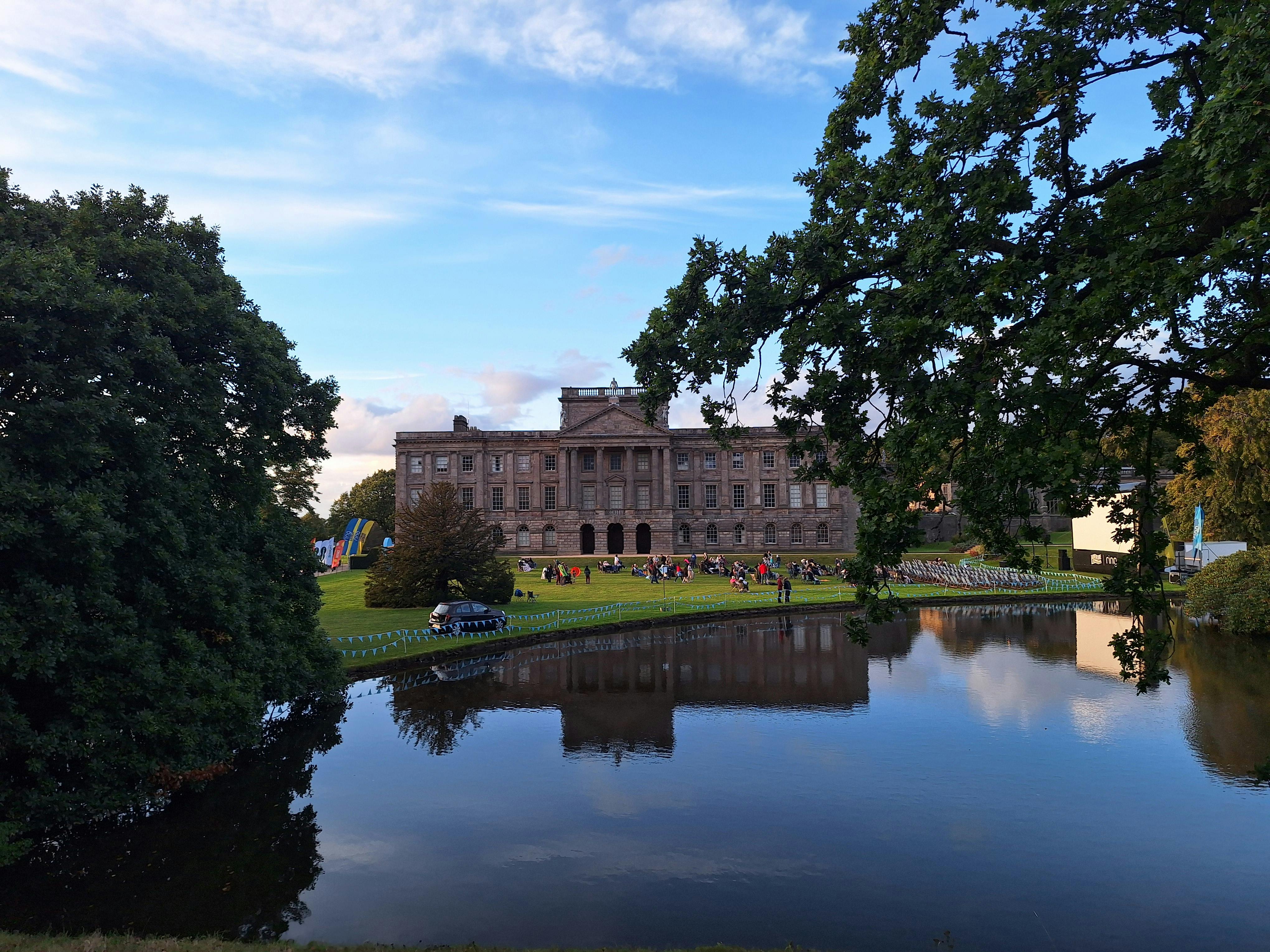 A grand English manor reflected in a tranquil pond with lush surroundings under a bright sky.