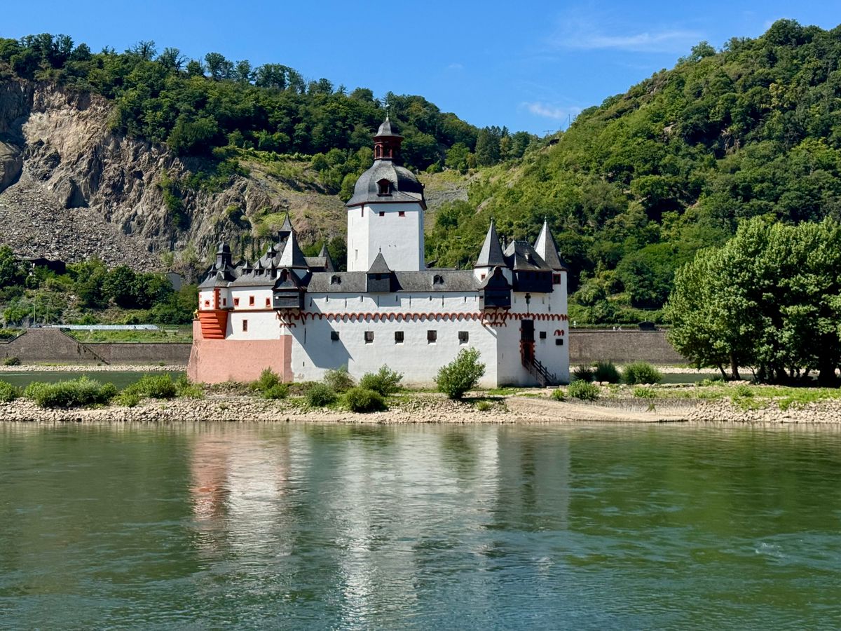 A white castle with grey-roofed towers and a red-pink base on a river island ringed with pale stones, with a bare rock face and green hills behind