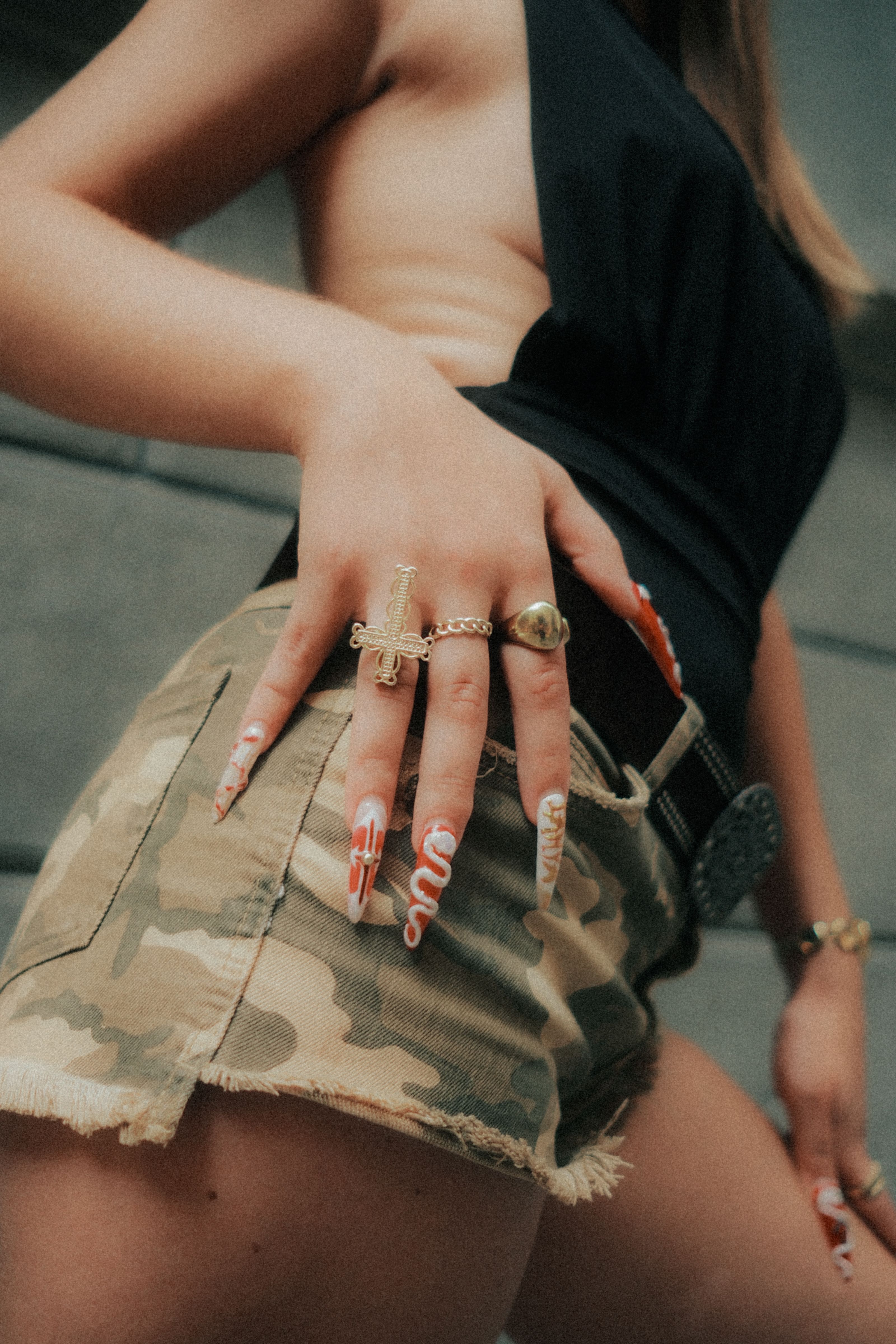 Close-up da mão de uma mulher com unhas acrílicas longas e anéis, vestindo shorts camuflados.
