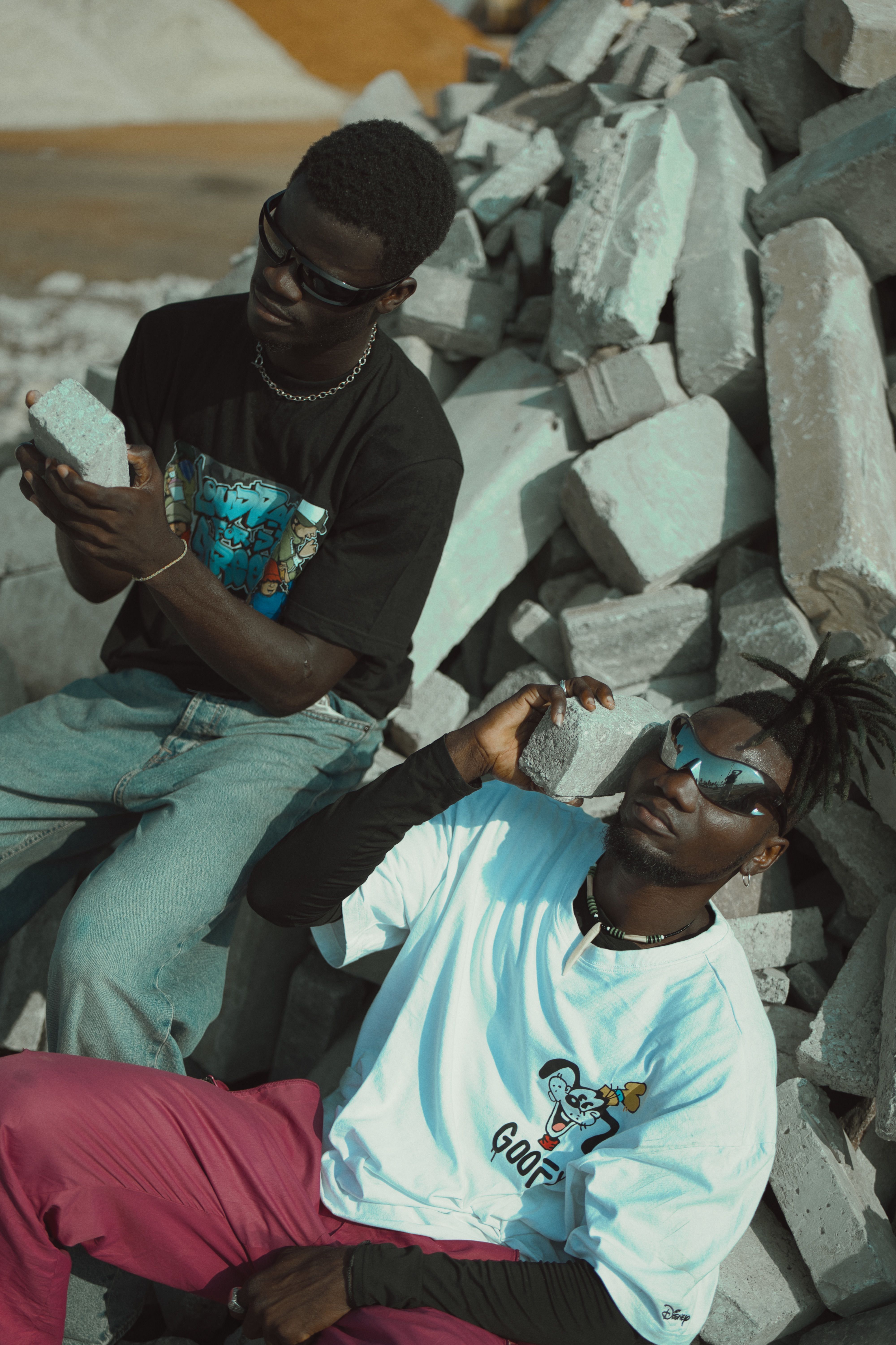 Two men in stylish attire pose among concrete rubble, showcasing urban fashion.