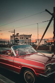 A classic red convertible parked near a pier at sunset with a warm glowing sky.