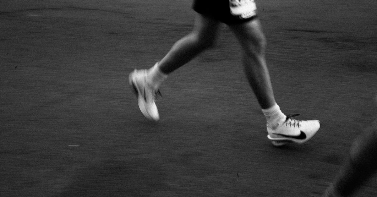 A black-and-white motion-blur close-up of a runner's legs striding across a city street, pure speed in asphalt and near-black tones.