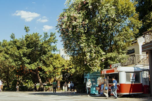 A lively scene with a coffee kiosk amidst lush trees and people enjoying a sunny day outdoors.