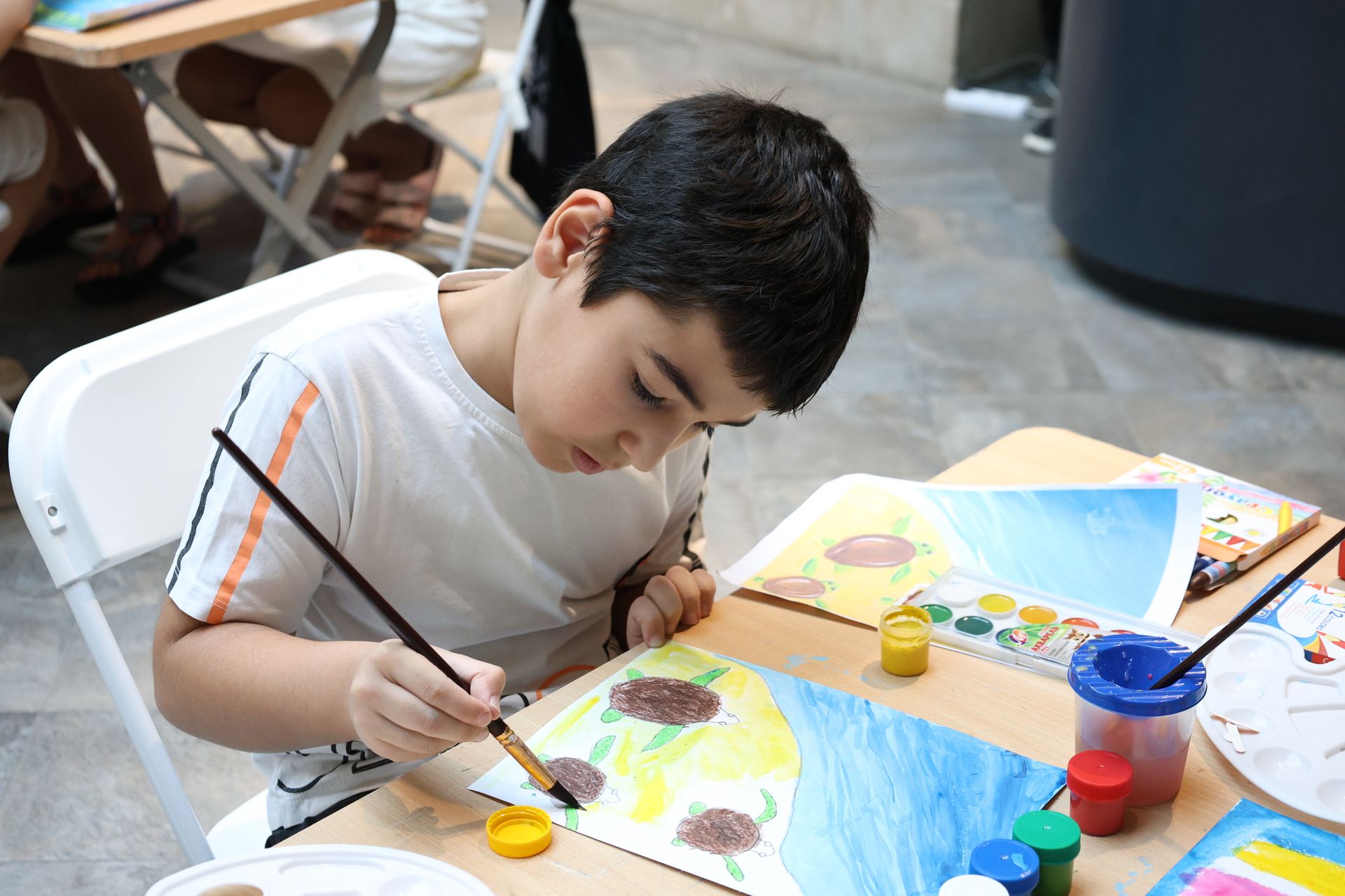 Child focused on painting at a table with colorful paints and brushes at an indoor art class.