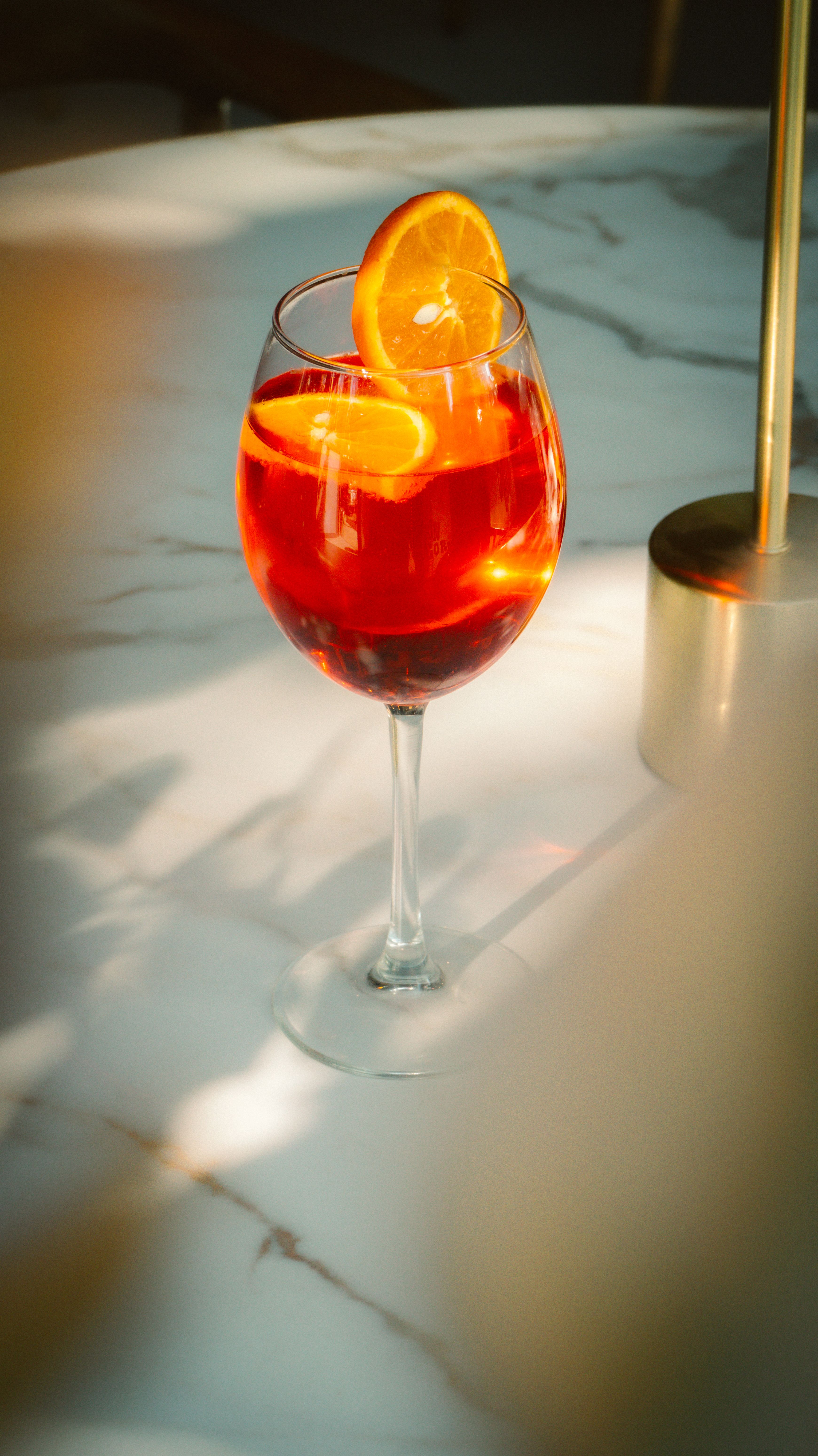 A glass of vibrant citrus sangria with orange slices on a marble table in sunlight.