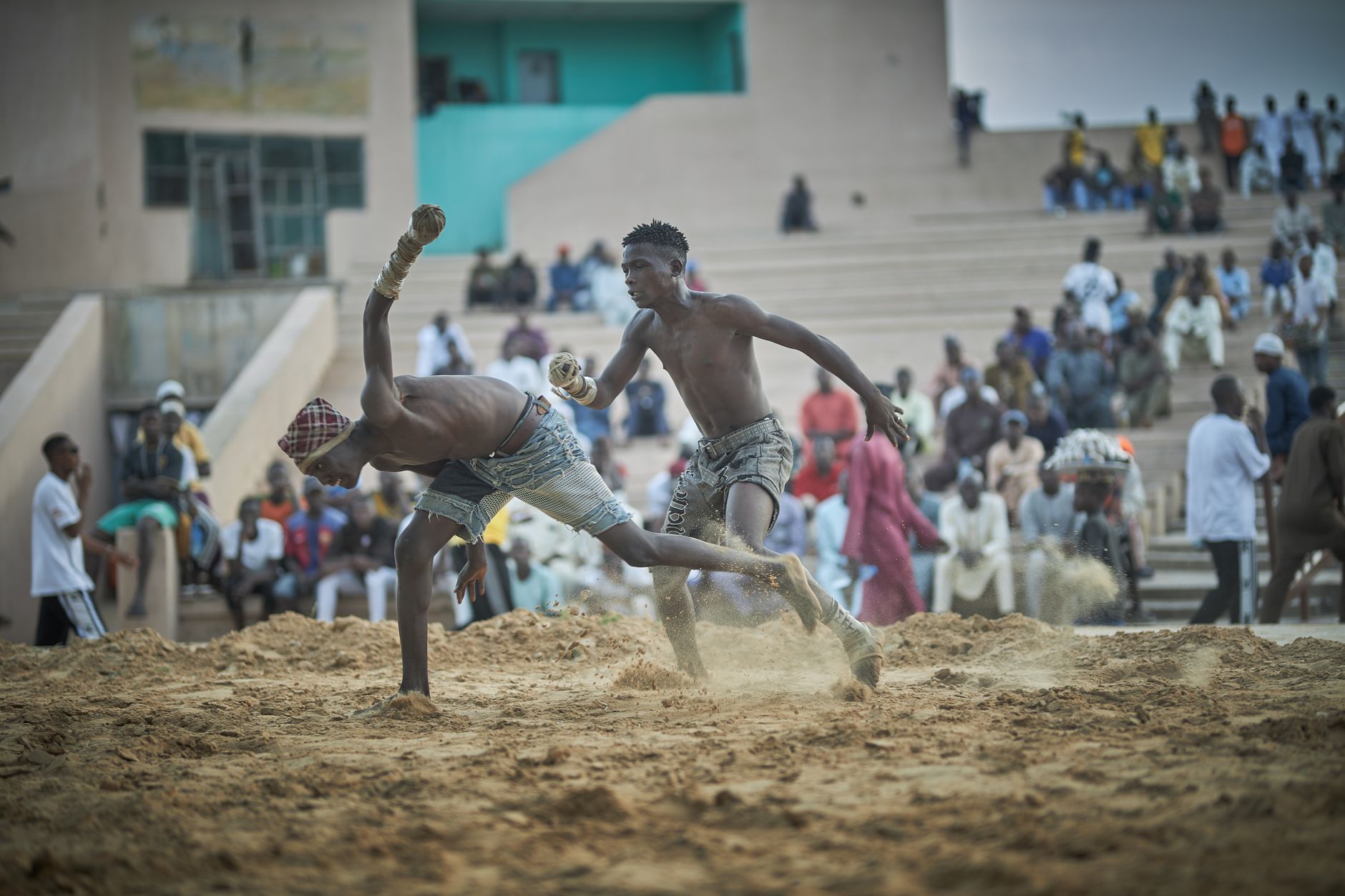 Action-packed scene of two men wrestling in a sandy arena with spectators.