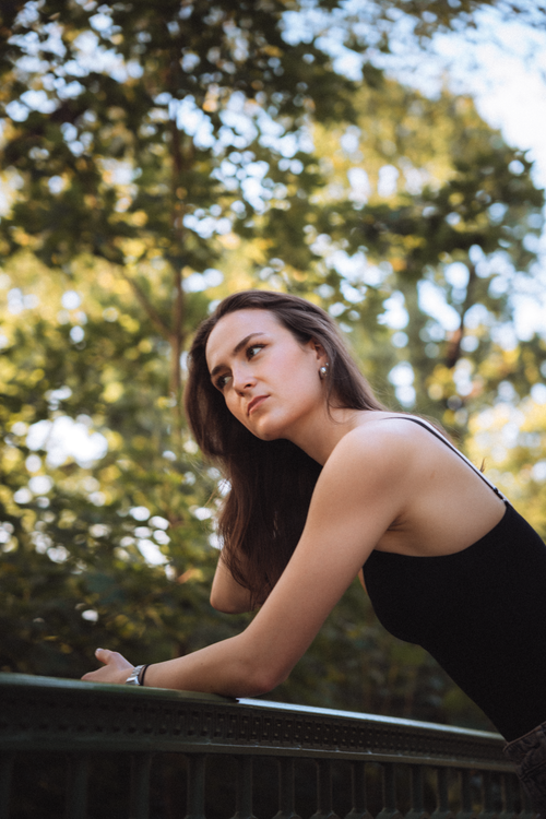 Free Young woman in a black dress leaning on a bridge railing in a park with trees. Stock Photo