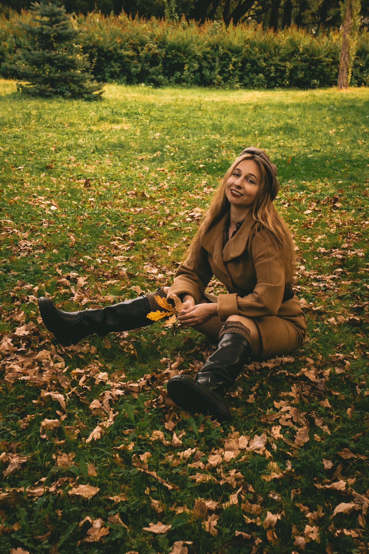 Cheerful woman in autumn attire sitting on a grassy lawn covered with fallen leaves, embracing the season.