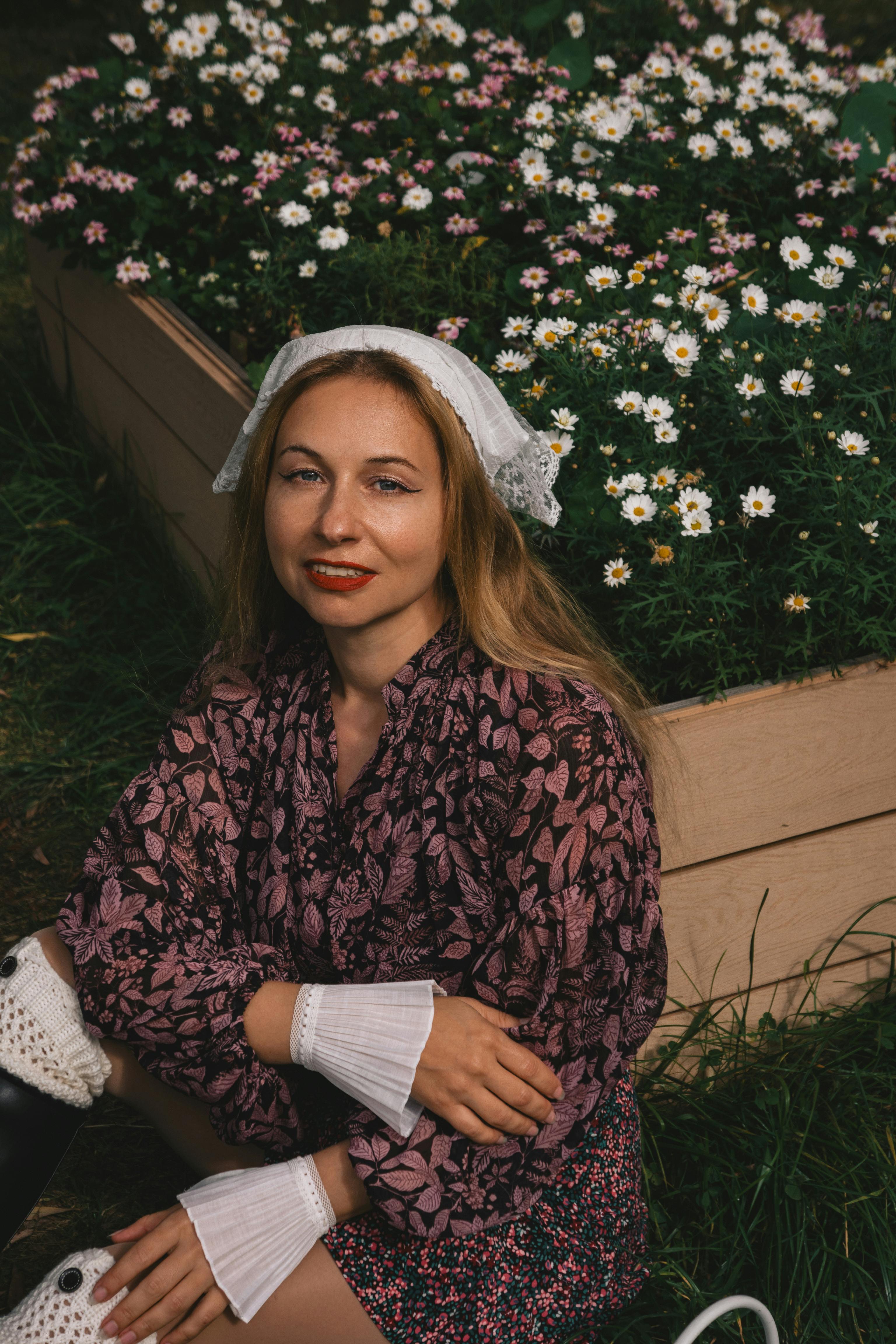 Smiling woman in garden with daisies, wearing floral attire and headscarf.