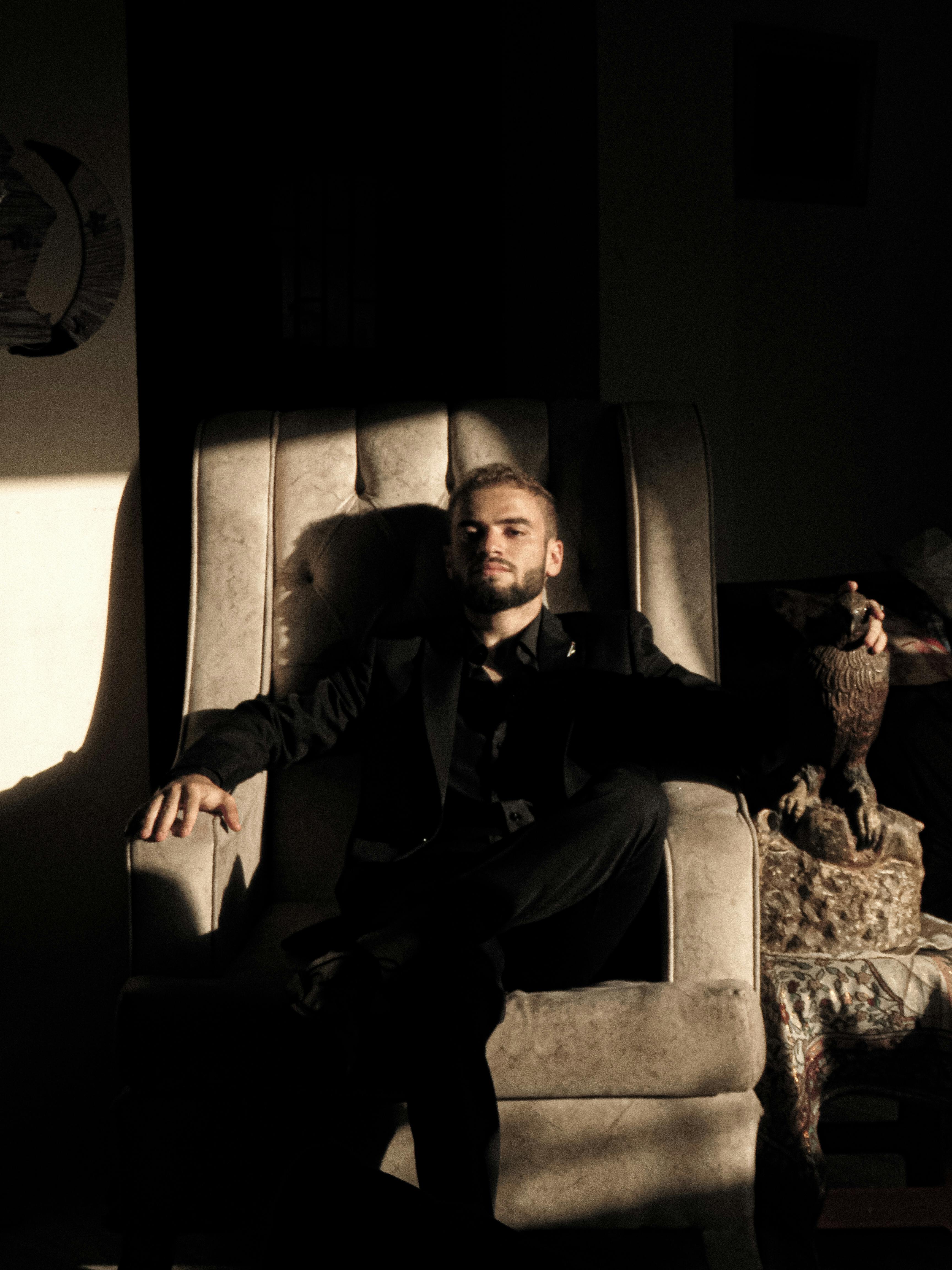 Gratuit Portret sumbru al unui bărbat așezat într-un fotoliu, proiectând umbre adânci în lumina blândă. Fotografie de stoc