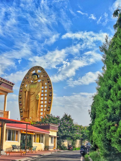 Free A towering Buddha statue stands under a bright blue sky, surrounded by lush greenery and architecture. Stock Photo