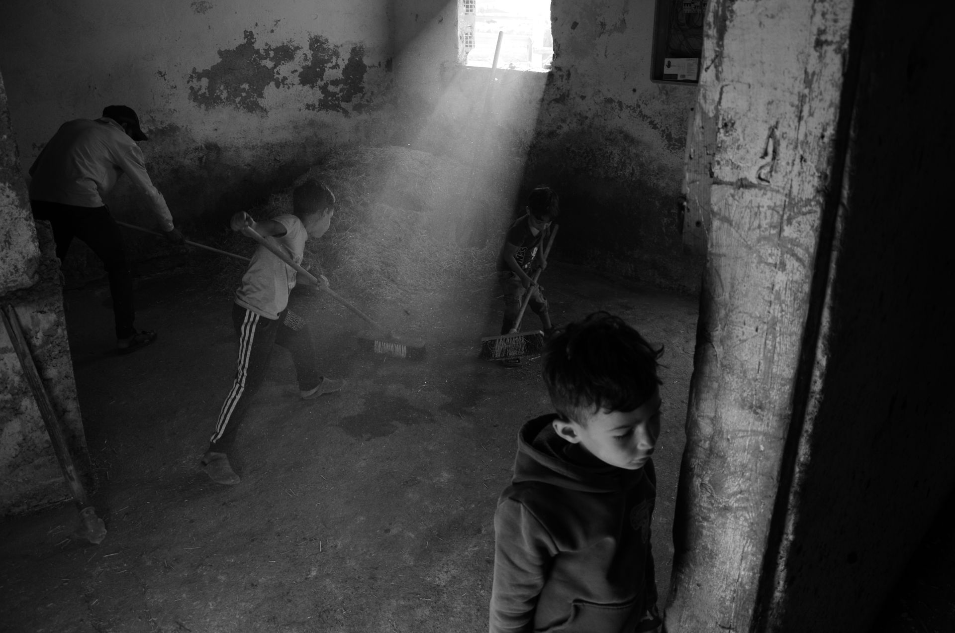 Black and white photo of children sweeping in a dusty room with light streaming through a window.