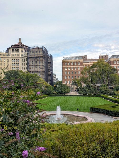 Free Serene park scene with a fountain, lush greenery, and historic buildings in the background. Stock Photo