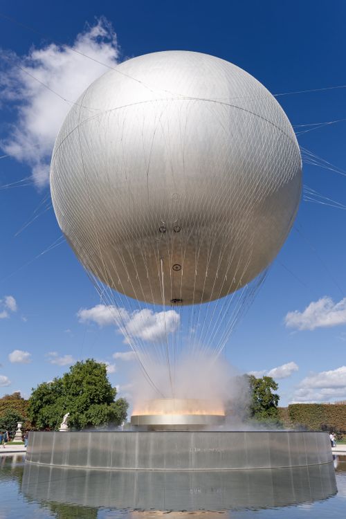 Free A large silver balloon soaring above a dazzling fountain in a Paris park under a bright blue sky. Stock Photo