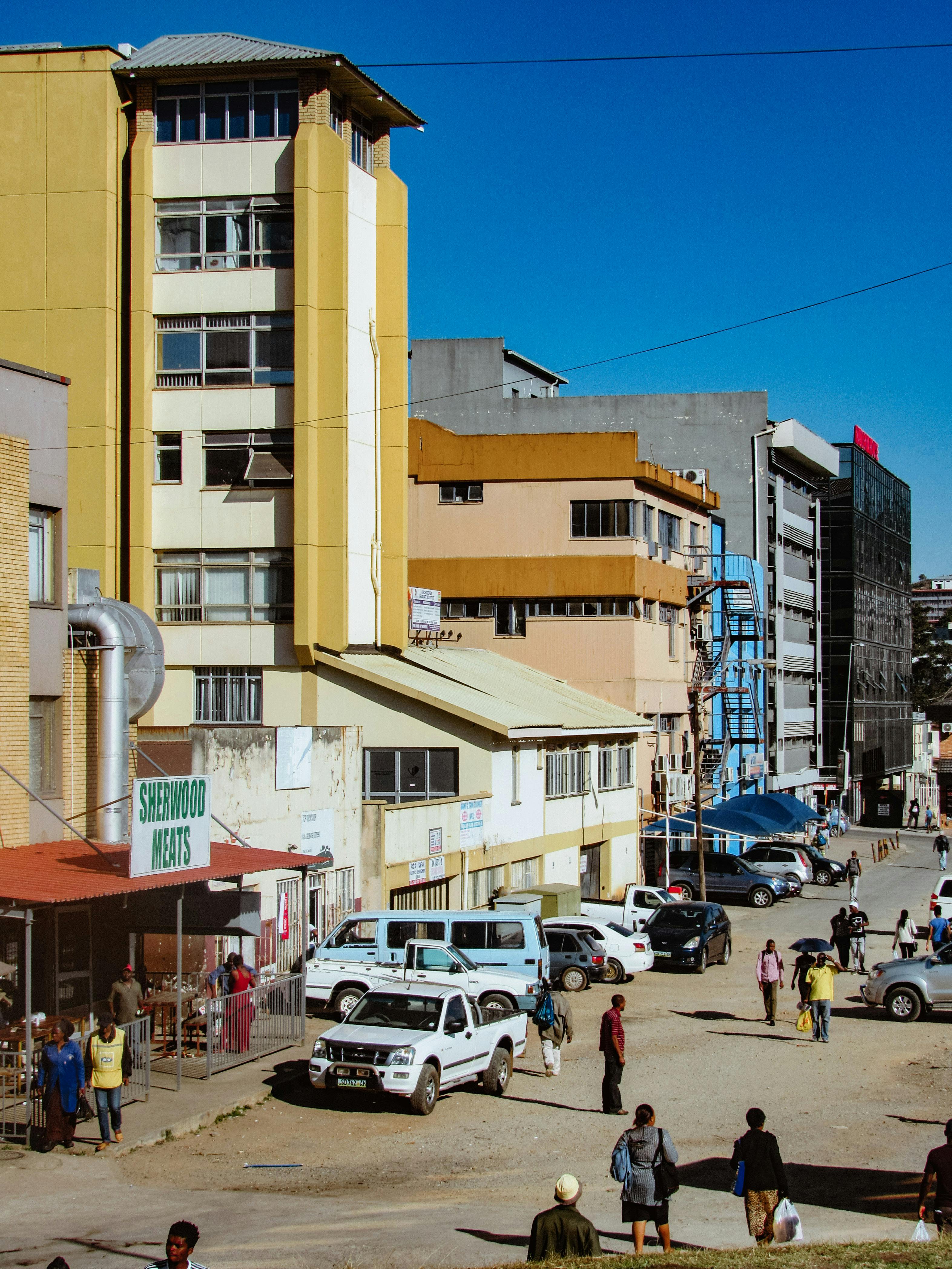 A vibrant urban street scene in Manzini, Eswatini with people and cars in the afternoon sun.