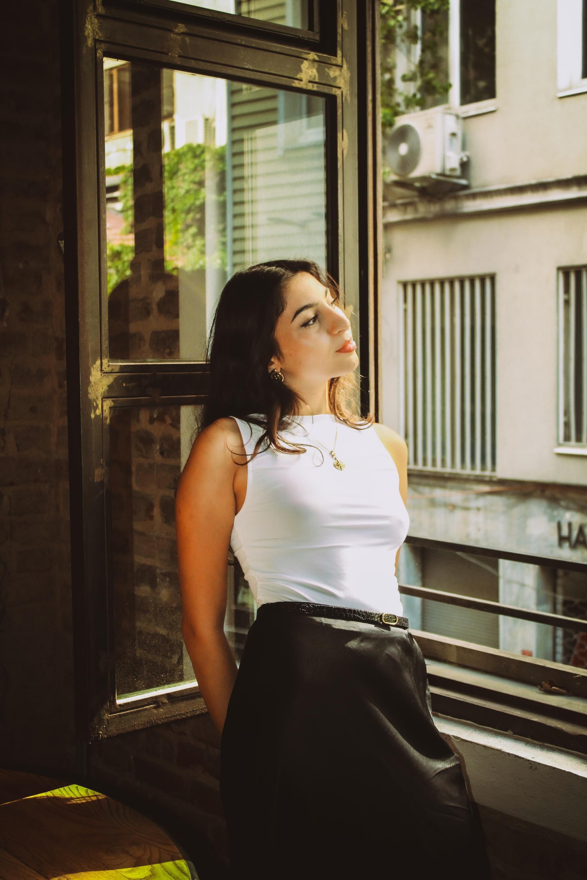 Woman in white top leans by an open window, bathed in soft light.