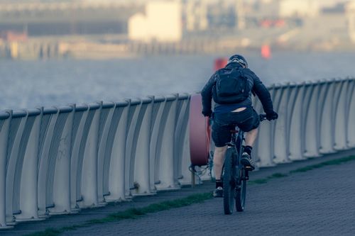 Free Cyclist with backpack rides along a waterfront path. Urban landscape in the background. Stock Photo