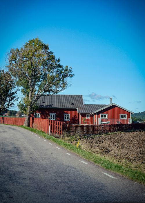 Free A scenic view of a red farmhouse with a tree-lined road under a clear blue sky. Stock Photo