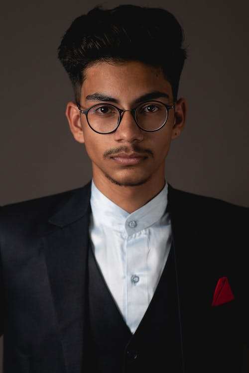 Free Portrait of stylish young Indian male model in formal suit and eyeglasses looking at camera against gray background Stock Photo