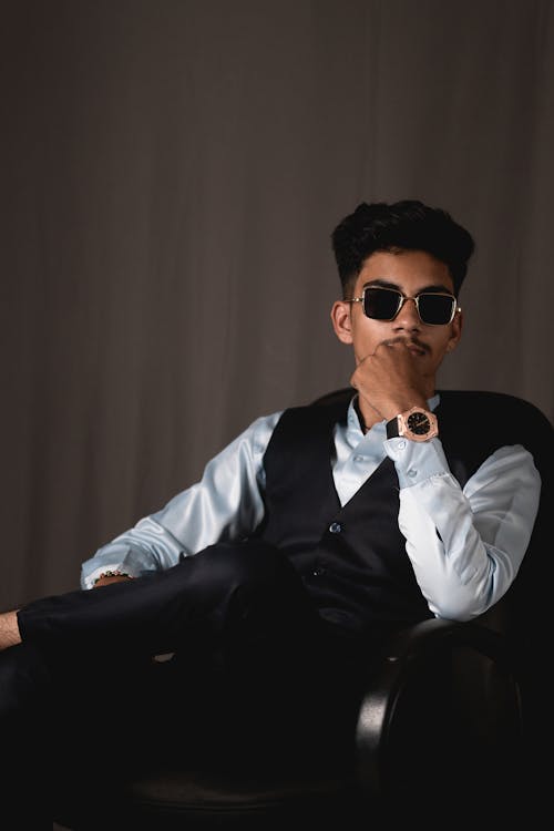 Serious young ethnic male manager in stylish formal suit and sunglasses sitting in comfortable armchair and touching chin