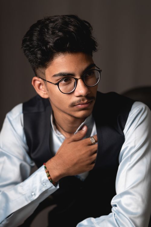 Free Portrait of concentrated young Indian male employee in formal outfit and eyeglasses sitting on chair and looking away thoughtfully against gray background Stock Photo
