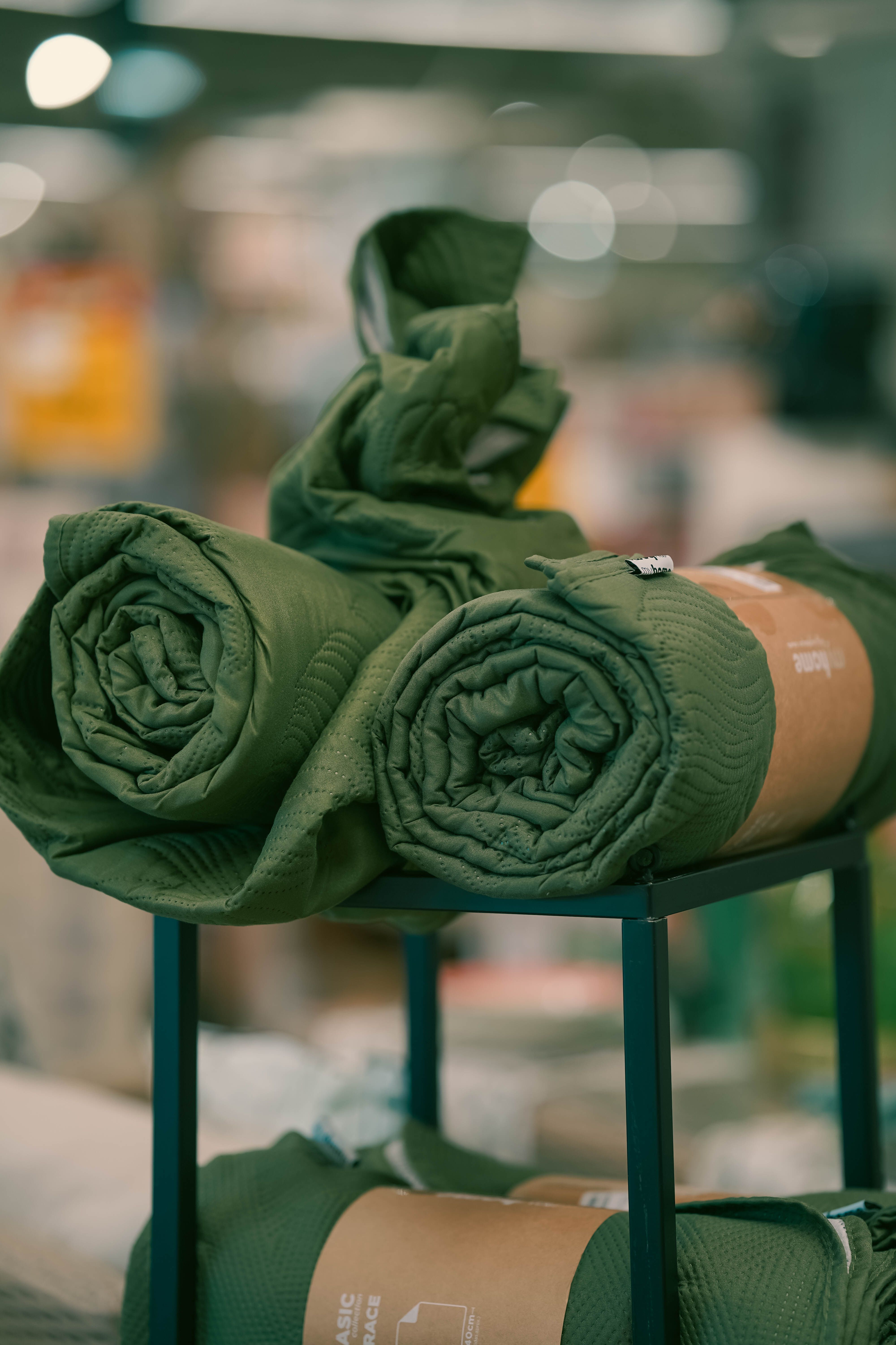 Three rolled green fabrics neatly displayed on a black stand indoors, with a blurred background.