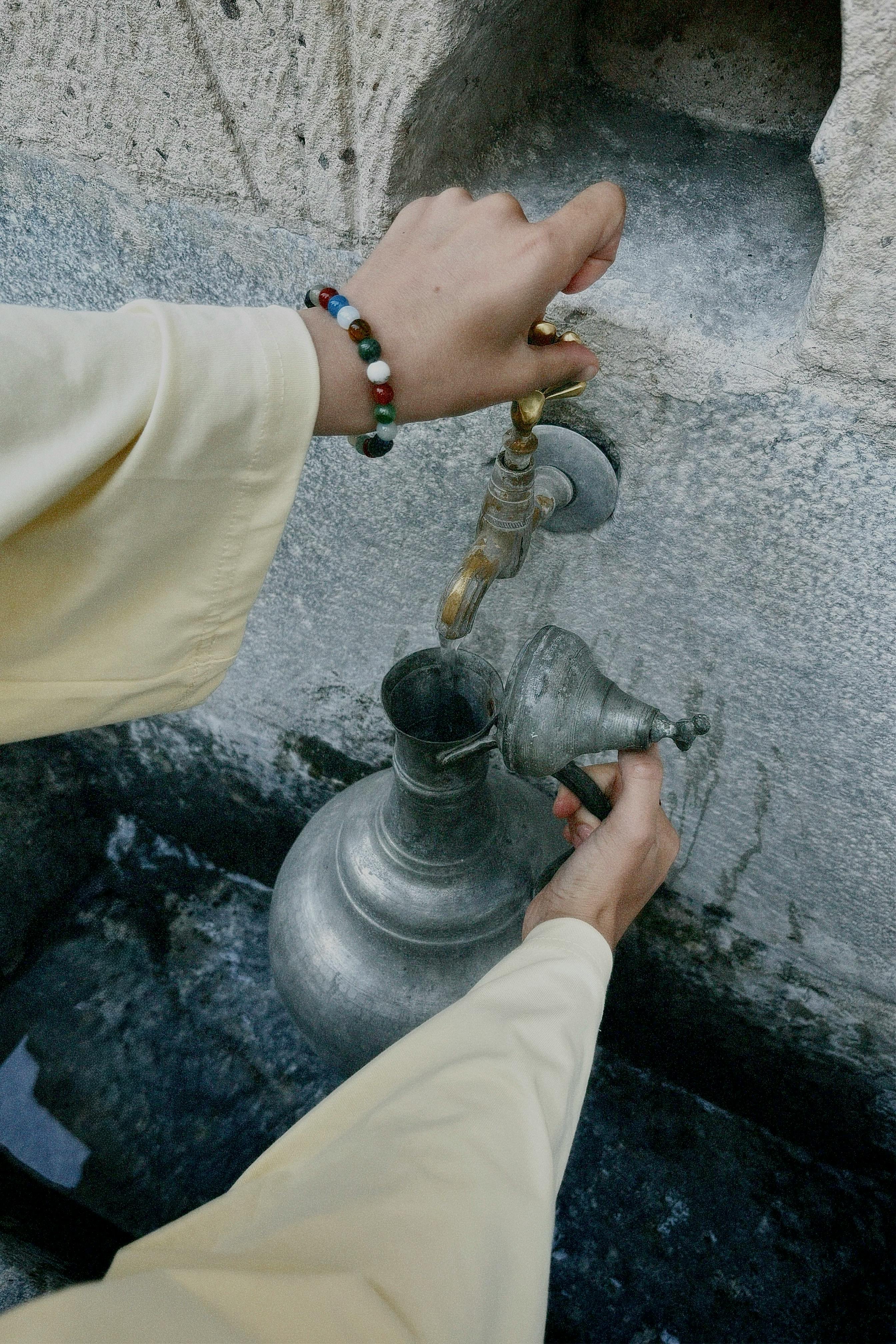 Hands filling a traditional metal jug with water from an outdoor stone faucet.