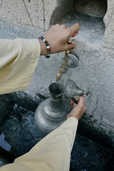 Hands filling a traditional metal jug with water from an outdoor stone faucet.