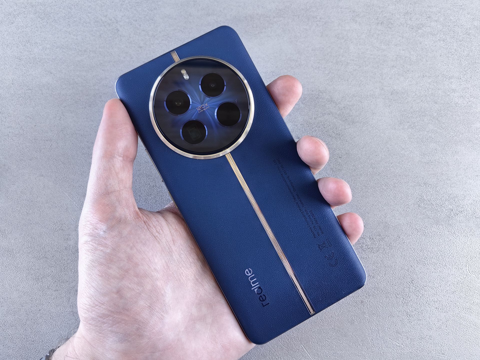 Close-up of a hand holding a blue smartphone featuring a triple camera design on a gray background.