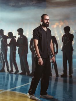 A basketball coach poses confidently indoors with his team silhouetted in a gym in Baghdad, Iraq.