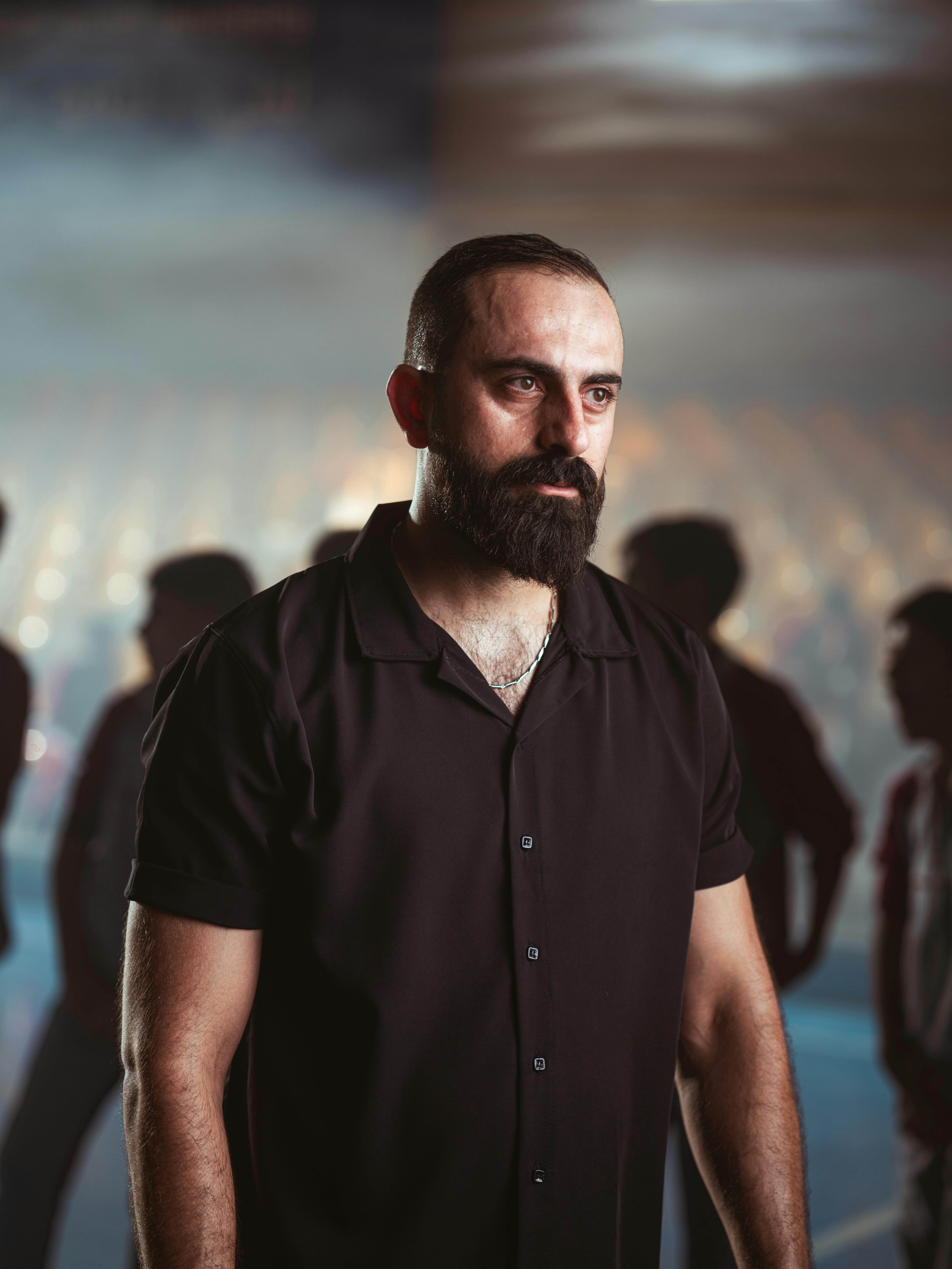 Ücretsiz Ciddi görünümlü sakallı bir adam dramatik bir ışık altında duruyor, yoğunluk ve odaklanmayı yansıtıyor. Stok Fotoğraflar