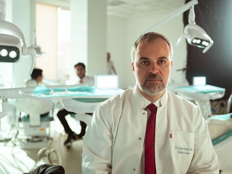 A bearded dentist in a white coat and red tie seated in a clinic