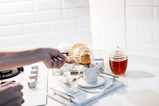 Hand pouring coffee into a mug in a bright, modern kitchen with honey and sugar on the side.