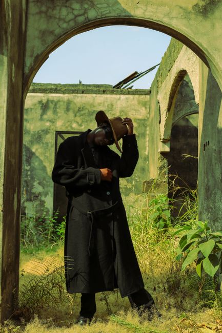 Man in a coat and hat in an abandoned, overgrown structure, striking a dramatic pose.