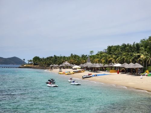 Free A serene tropical beach with jet skis, palm trees, and thatched umbrellas along the shoreline. Stock Photo