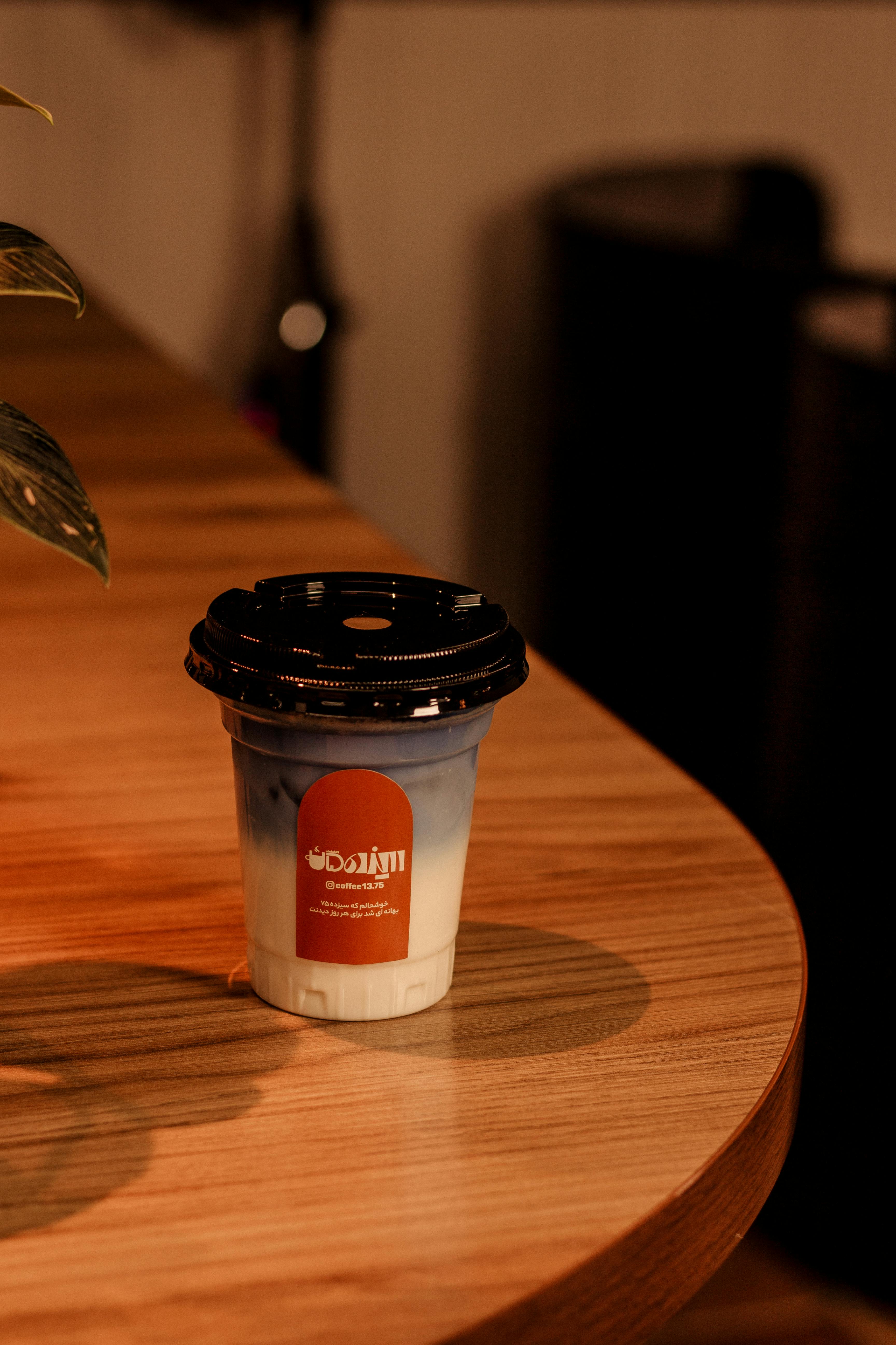 A warm coffee beverage in a takeaway cup on a wooden table at a cafe in Kermanshah, Iran.