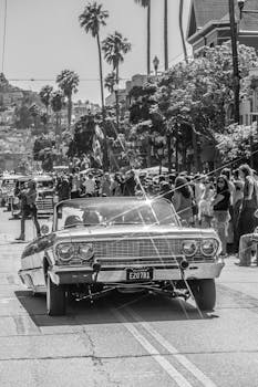 Black and white image of a classic convertible in a lively city street parade.