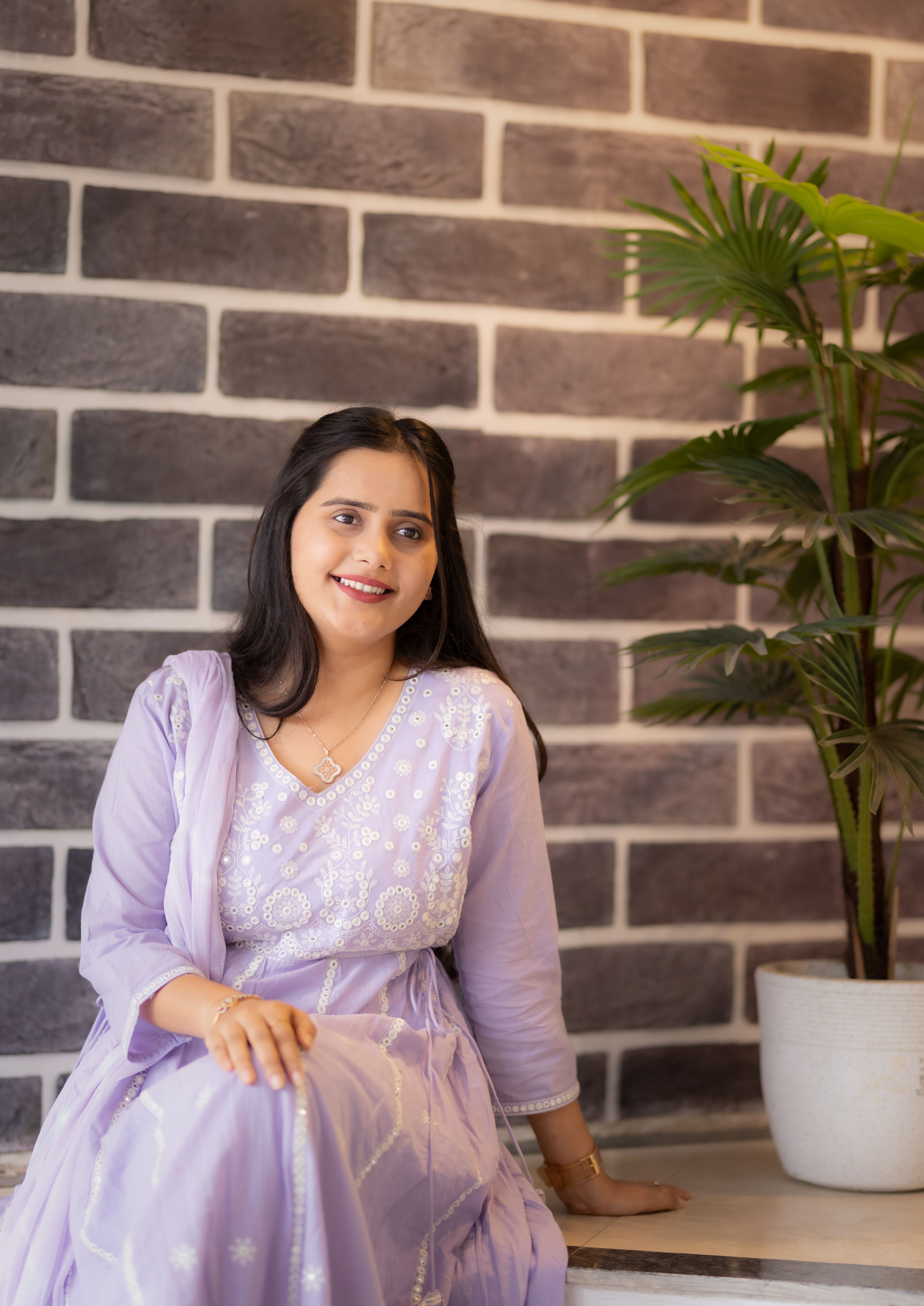 A graceful woman in lavender traditional Indian dress seated indoors next to a potted plant.