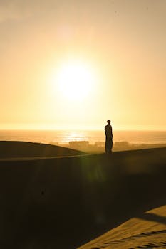 A lone figure walks on desert dunes at sunset, silhouetted against a vast, glowing sky.