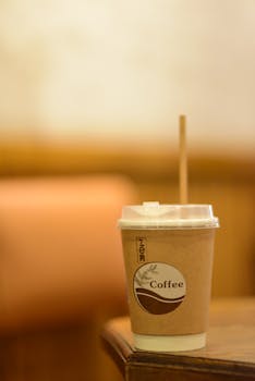 A close-up of a takeaway coffee cup with a wooden straw on a wooden table in a warm, cozy setting.