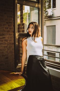 A fashionable woman in white top and black skirt stands by a sunlit window in an urban setting.