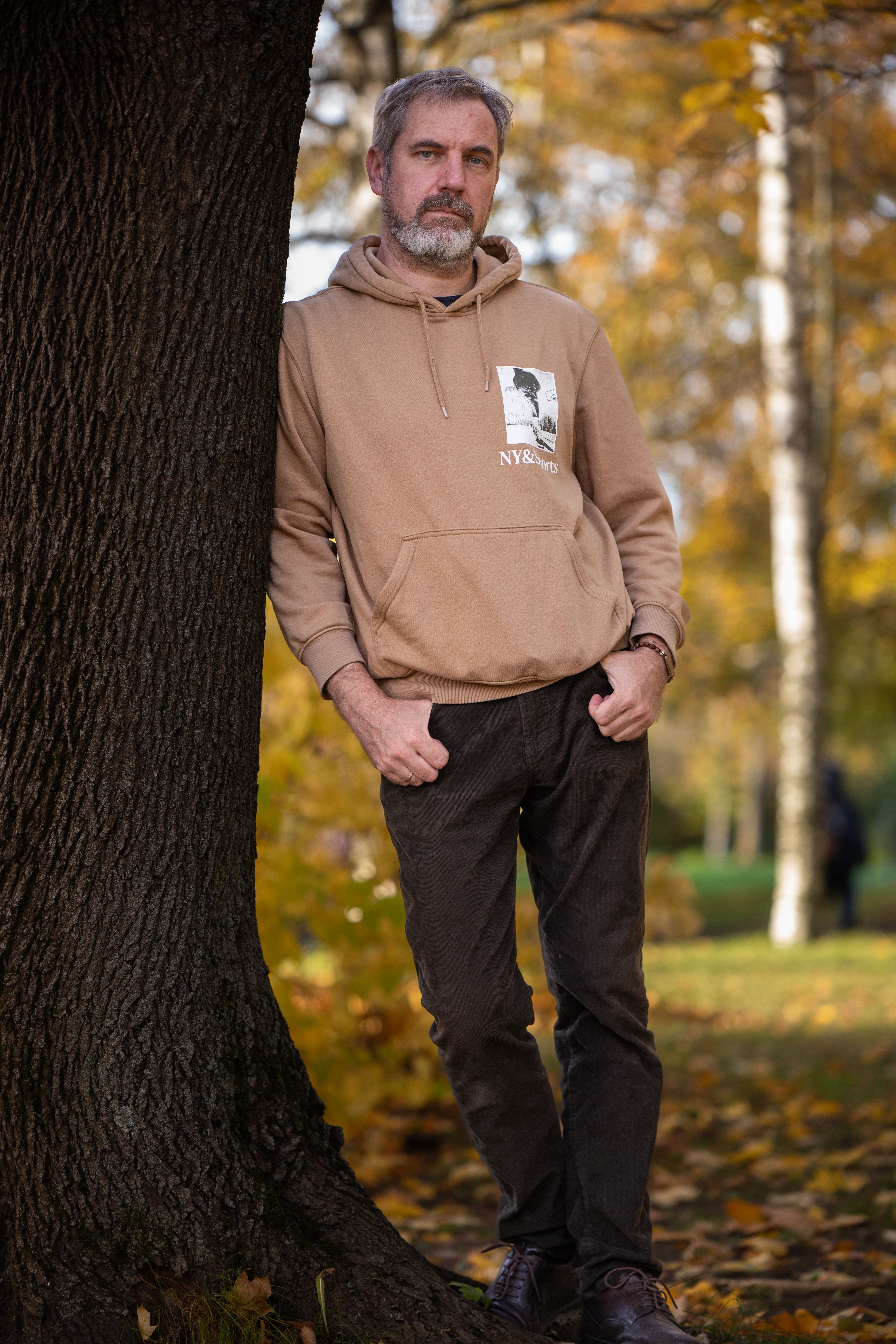 Casual man leans against tree in a park during fall, capturing a relaxed autumn vibe.